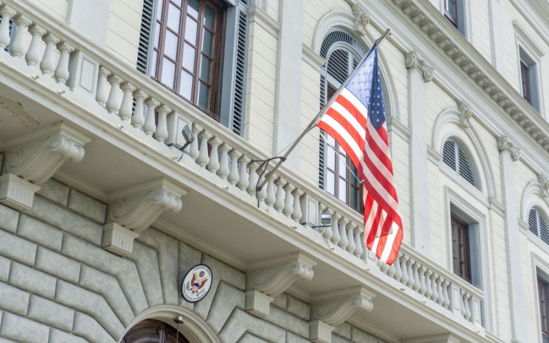 Mulher é presa após uso de explosivos em consulado dos EUA, imagem mostra fachada de prédio branco com bandeira americana pendurada